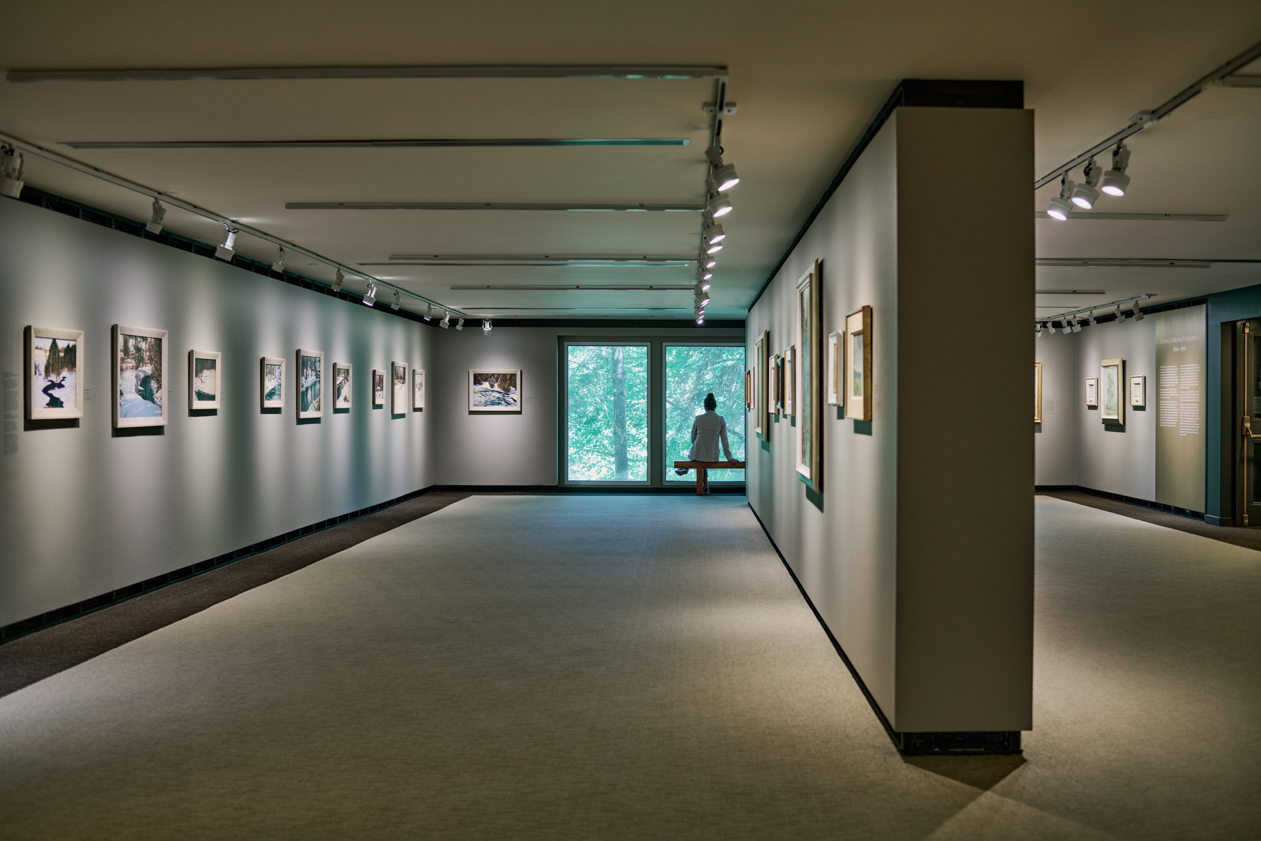 An art gallery with numerous paintings on the walls and a lone figure sitting on a bench looking out.