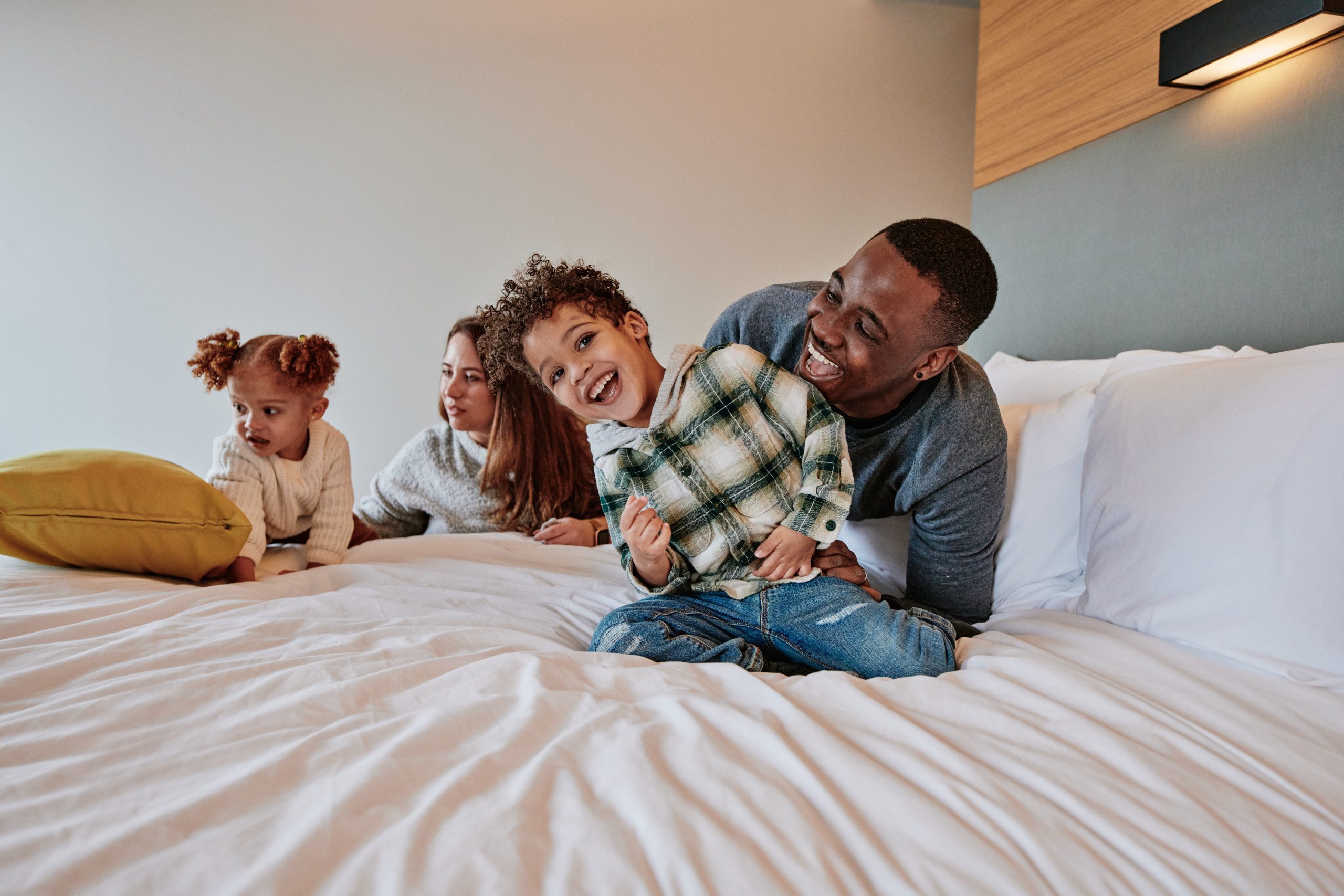 Two adults with two young children playing on a bed with white sheets.